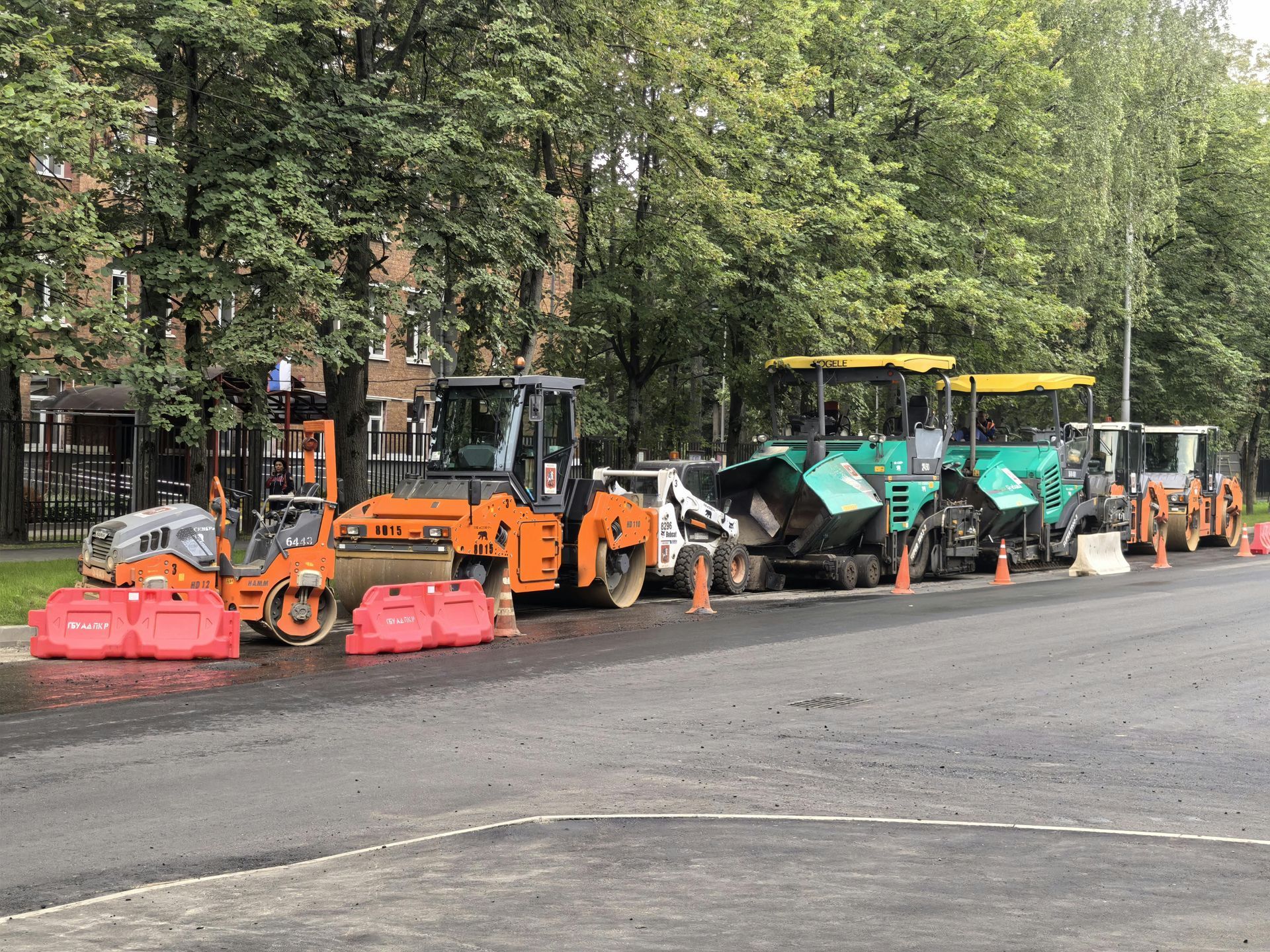 Road construction equipment lined up on a street with trees. Includes rollers, pavers, and cones.