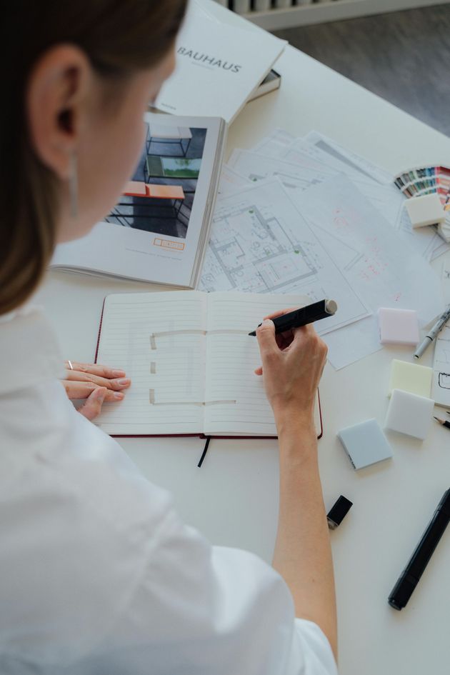 Person sketching a floor plan in a notebook on a white desk with architectural drawings and design materials.