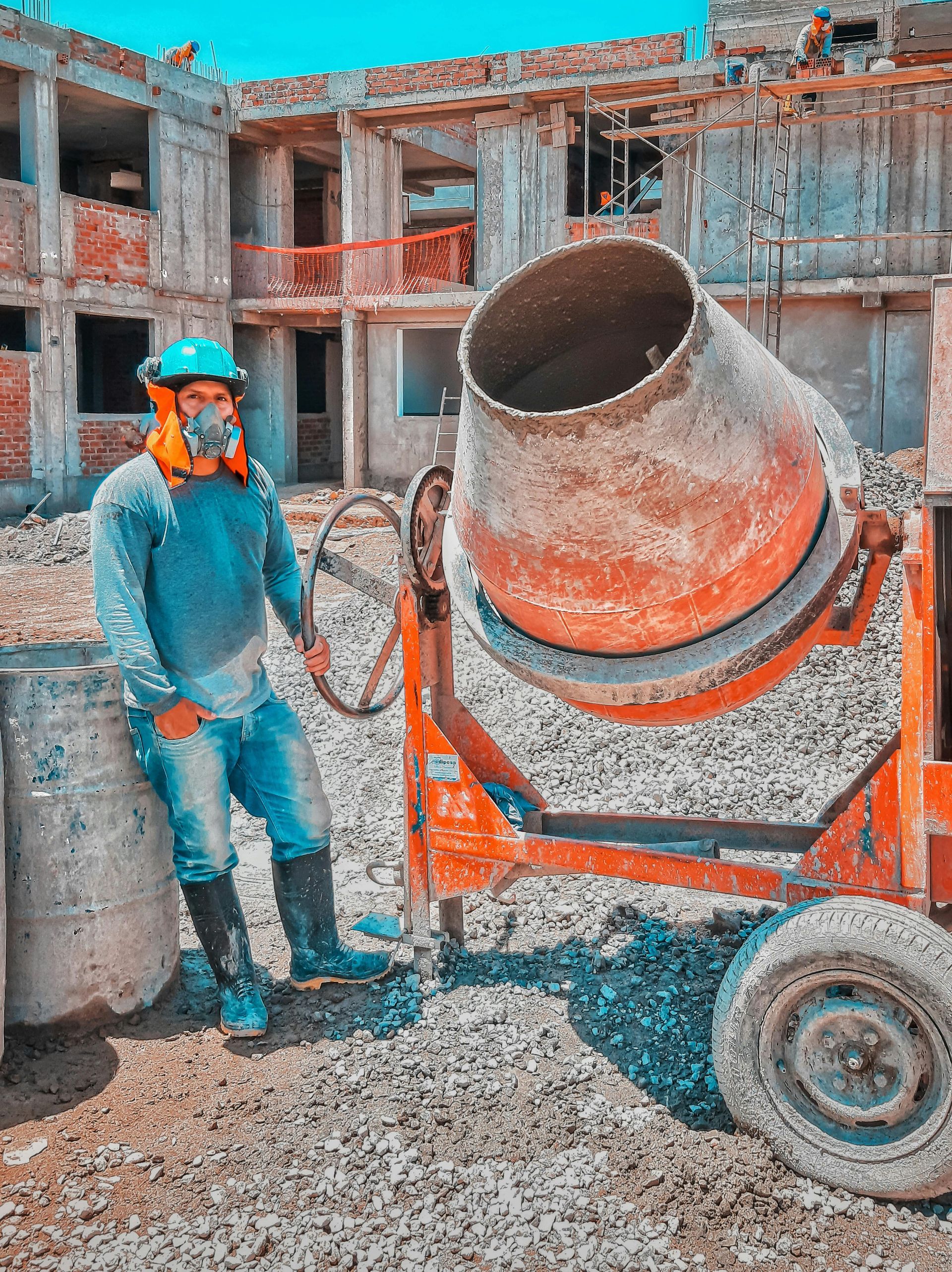 Construction worker beside an orange concrete mixer, wearing safety gear, at a building site.