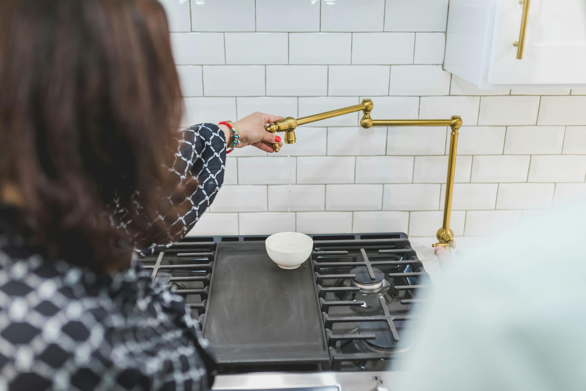 Person reaching for a pot filler faucet above a stovetop with a white bowl on it.