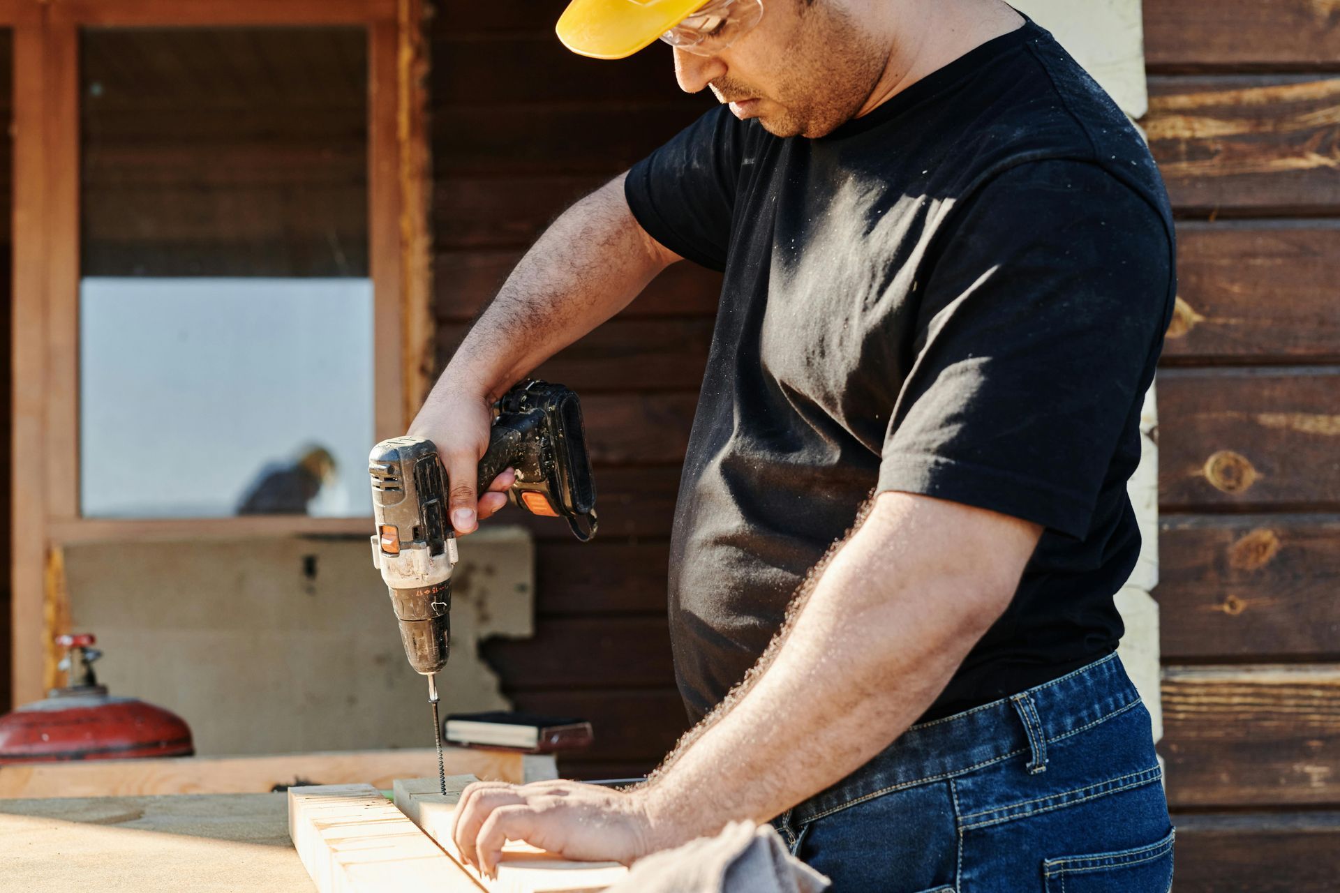 Man in black shirt and yellow hard hat using a drill on wood outdoors.