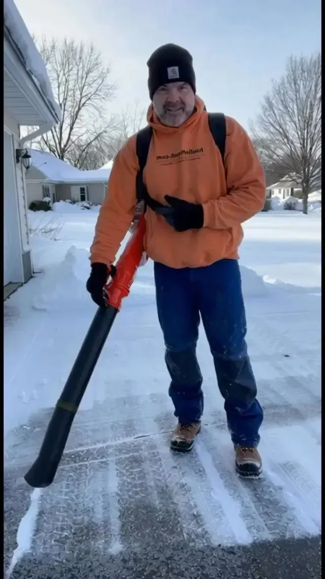 A person wearing a bright orange hoodie and black beanie stands in a snow-covered driveway, holding a leaf blower.