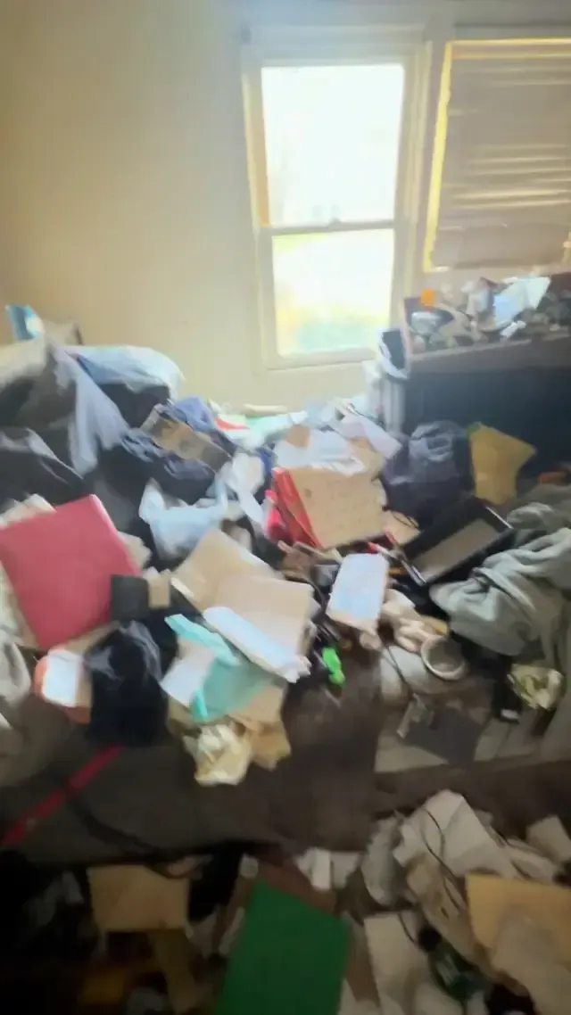 A cluttered room with a bed covered in piles of clothes, paper, and various belongings, illuminated by a nearby window.
