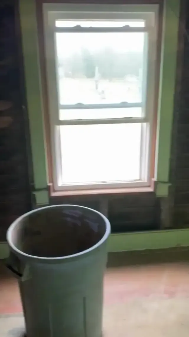 A light green trash can stands in an unfinished room with exposed brick walls and a single white double-hung window.
