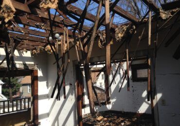 The charred interior of a room with exposed wooden roof beams, fire damage, and daylight visible through the open ceiling.