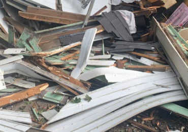 A pile of construction debris featuring white siding, wood scraps, and dark roofing shingles scattered on the ground.