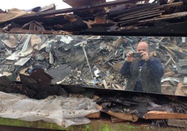 A person holding a phone to take a mirror selfie in front of a large pile of debris and building materials.