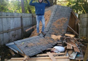 A person wearing a blue hoodie and jeans stands proudly on top of a collapsed wooden shed in a backyard.