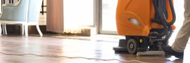 Orange floor-cleaning machine on a sunlit indoor floor near a chair and windows