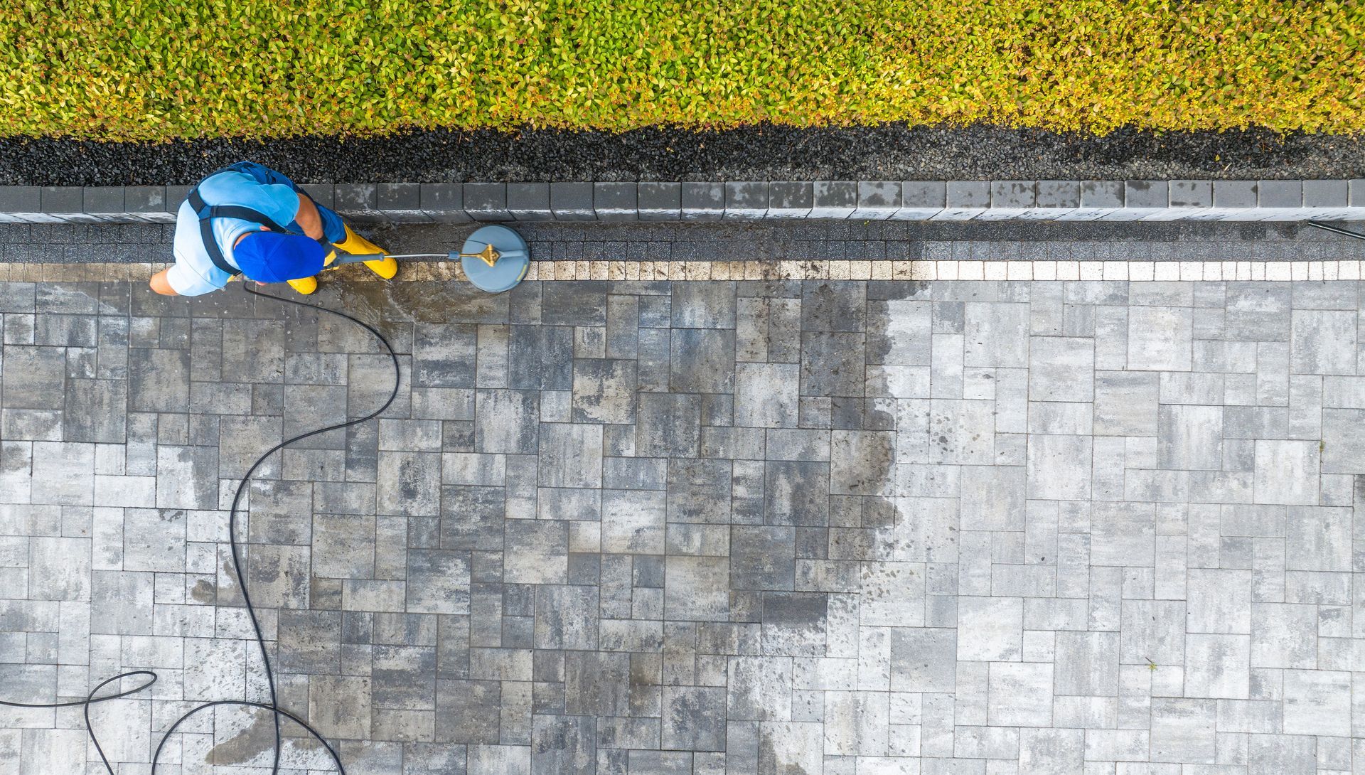 Worker in blue cleaning a stone sidewalk beside a curb with a hose and bucket.