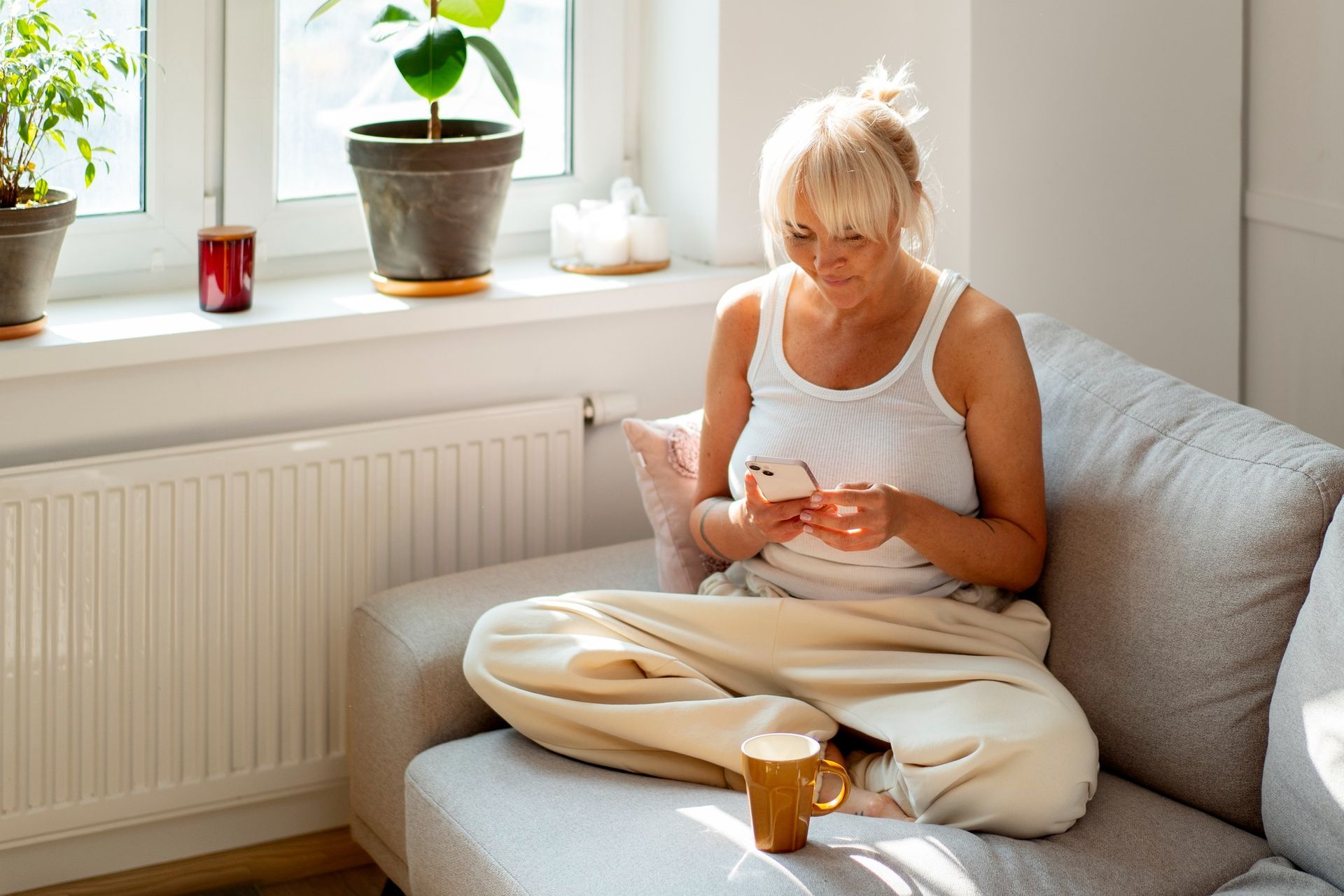 Person sitting on a gray sofa, looking at a phone, with a coffee mug and potted plant nearby.