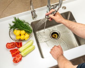 Hands washing vegetables at a kitchen sink; tomatoes, cucumbers, potatoes, and herbs on the counter