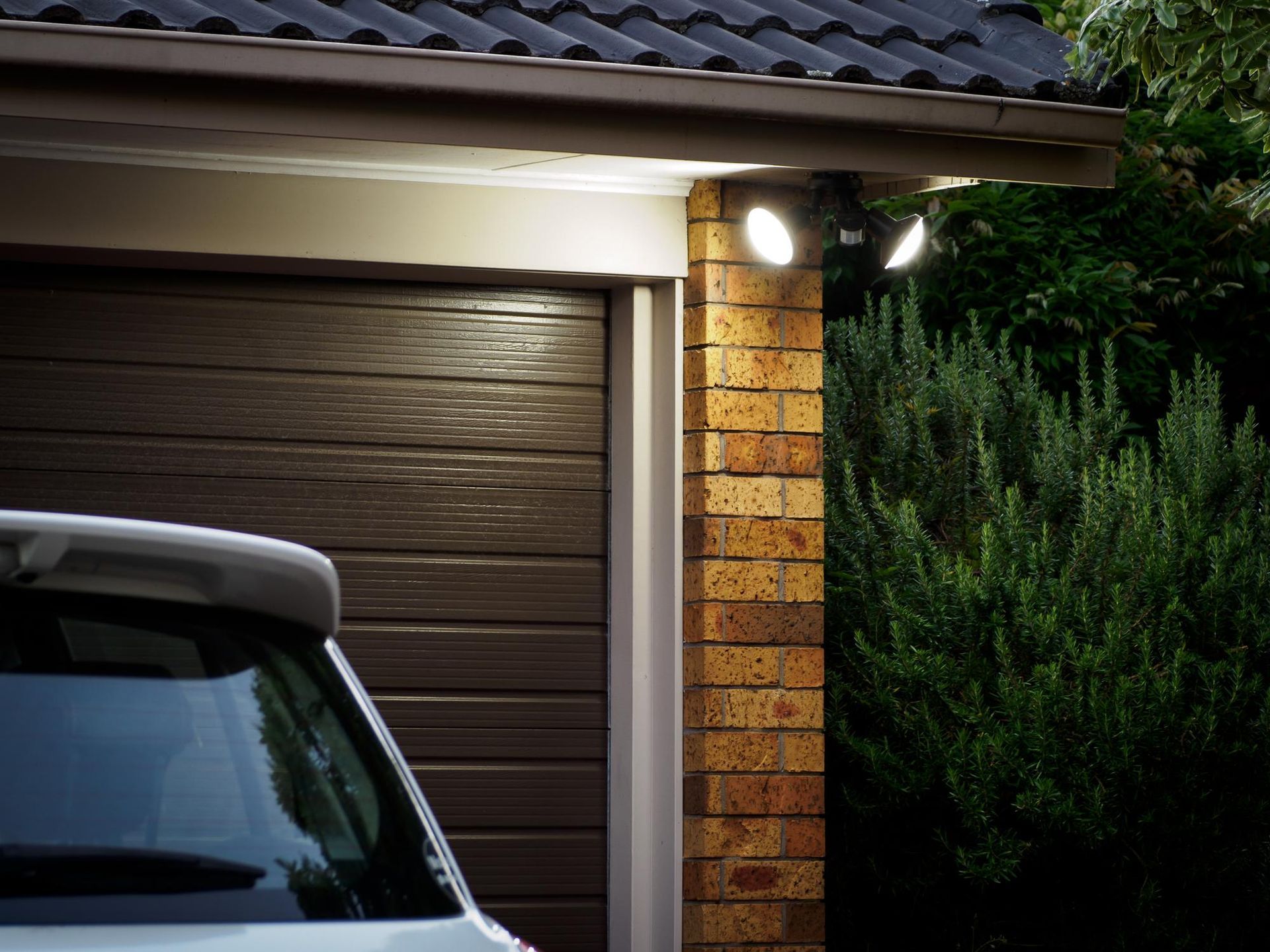 Exterior of a house garage with lit porch lights at dusk, partly showing a parked car and brick pillar