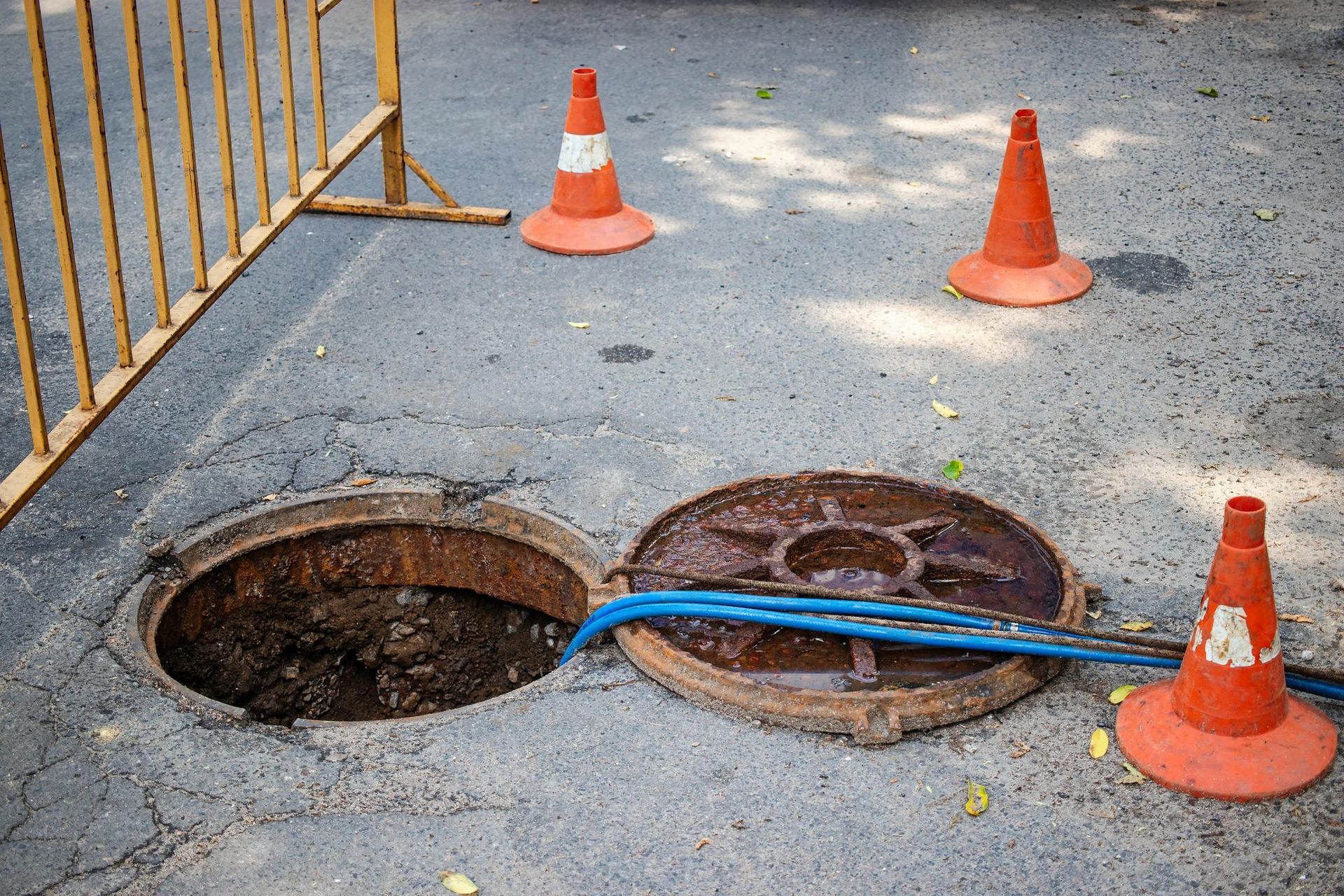 Open manhole and broken cover on pavement, marked by orange traffic cones and a blue hose