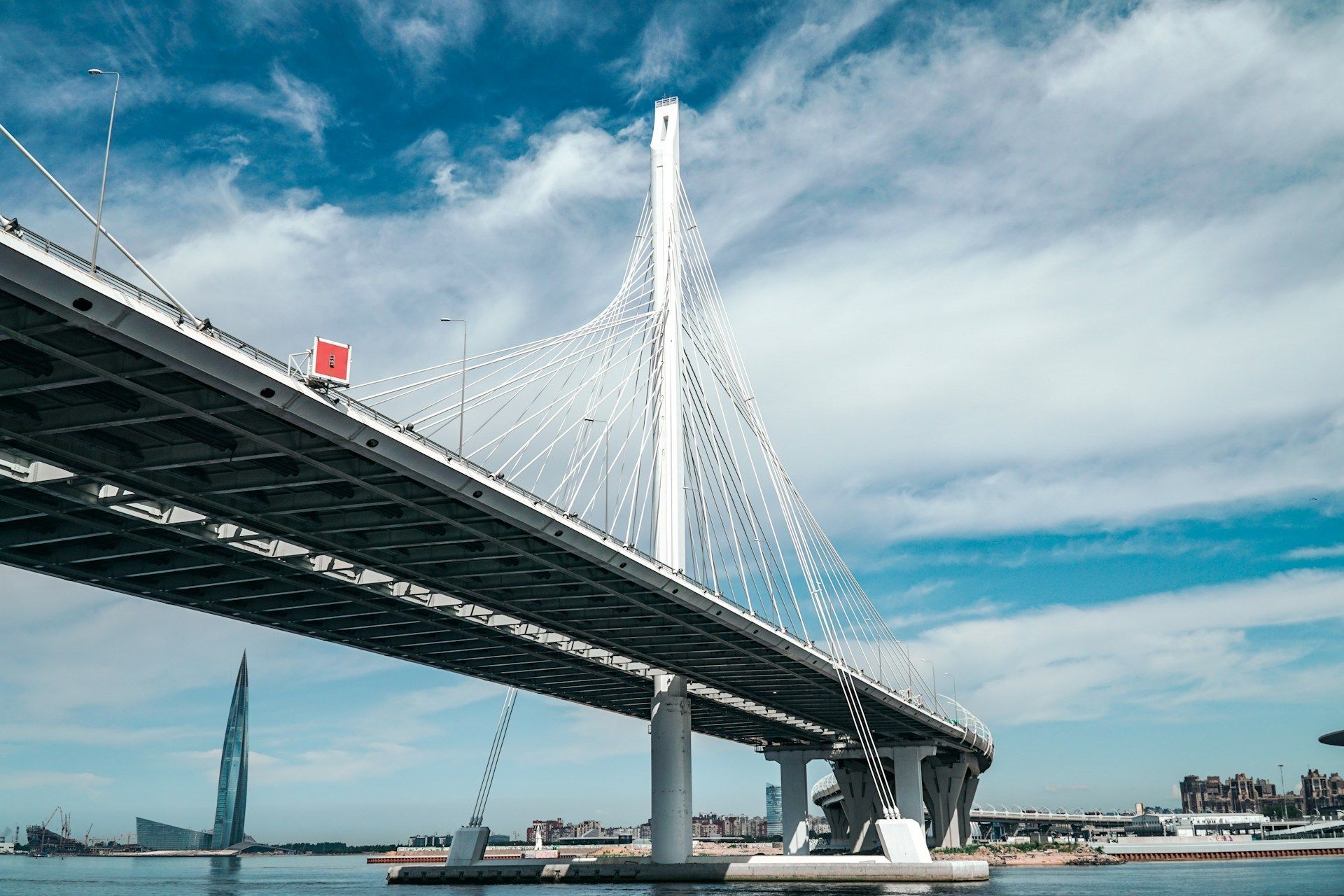 Cable-stayed bridge over water under a cloudy blue sky, with a roadway in the foreground