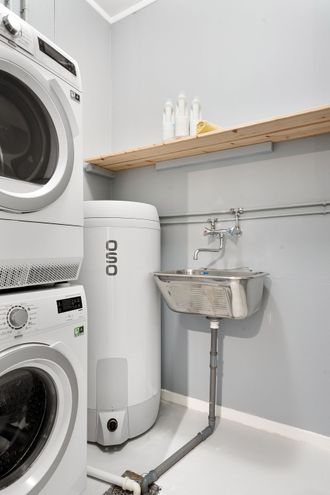Small laundry room with stacked washer-dryer, wall-mounted sink, and white water heater under a shelf.