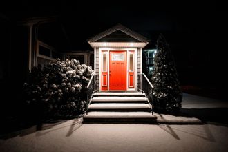 Red front door with white columns and steps, lit at night and flanked by bushes.
