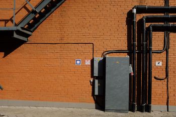 Orange brick wall with black pipes, a gray utility box, and a metal staircase on the left.