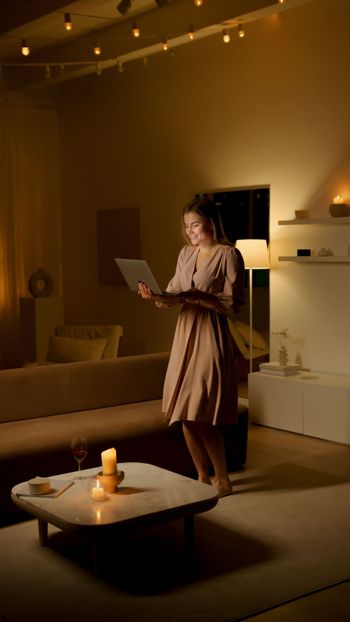 Woman standing in a cozy living room, reading from a tablet beside a coffee table and lamp-lit shelves