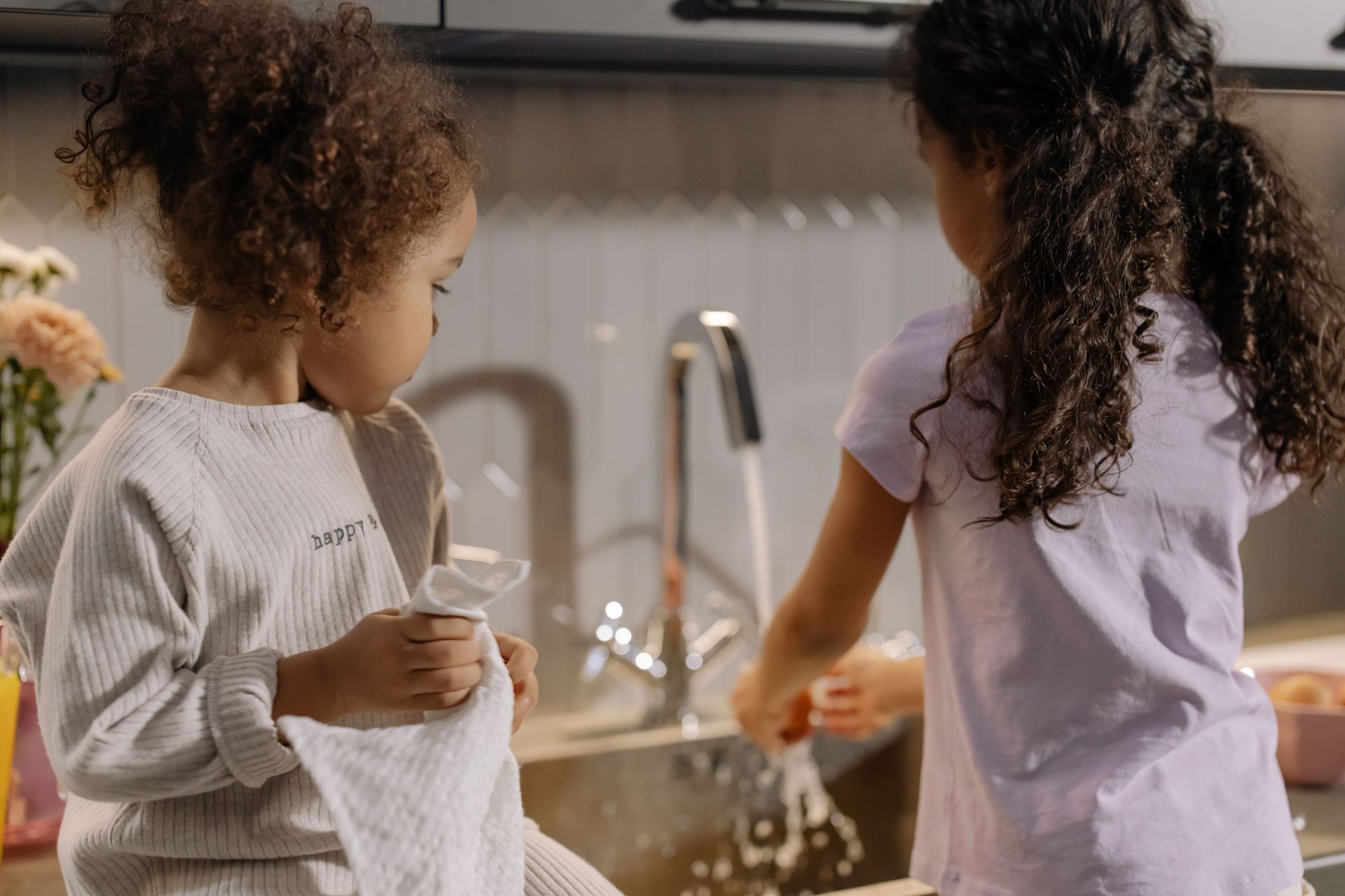 Two children standing at a kitchen sink, washing dishes with water running.