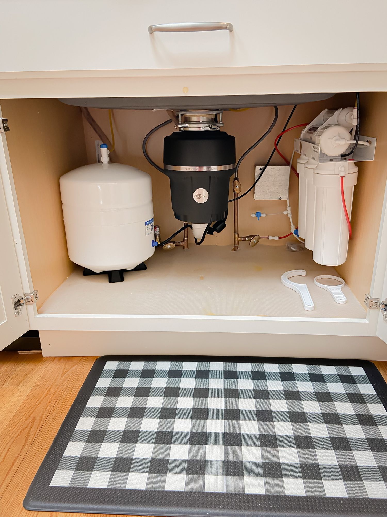 The underside of a kitchen sink with a black and white checkered mat on the floor.