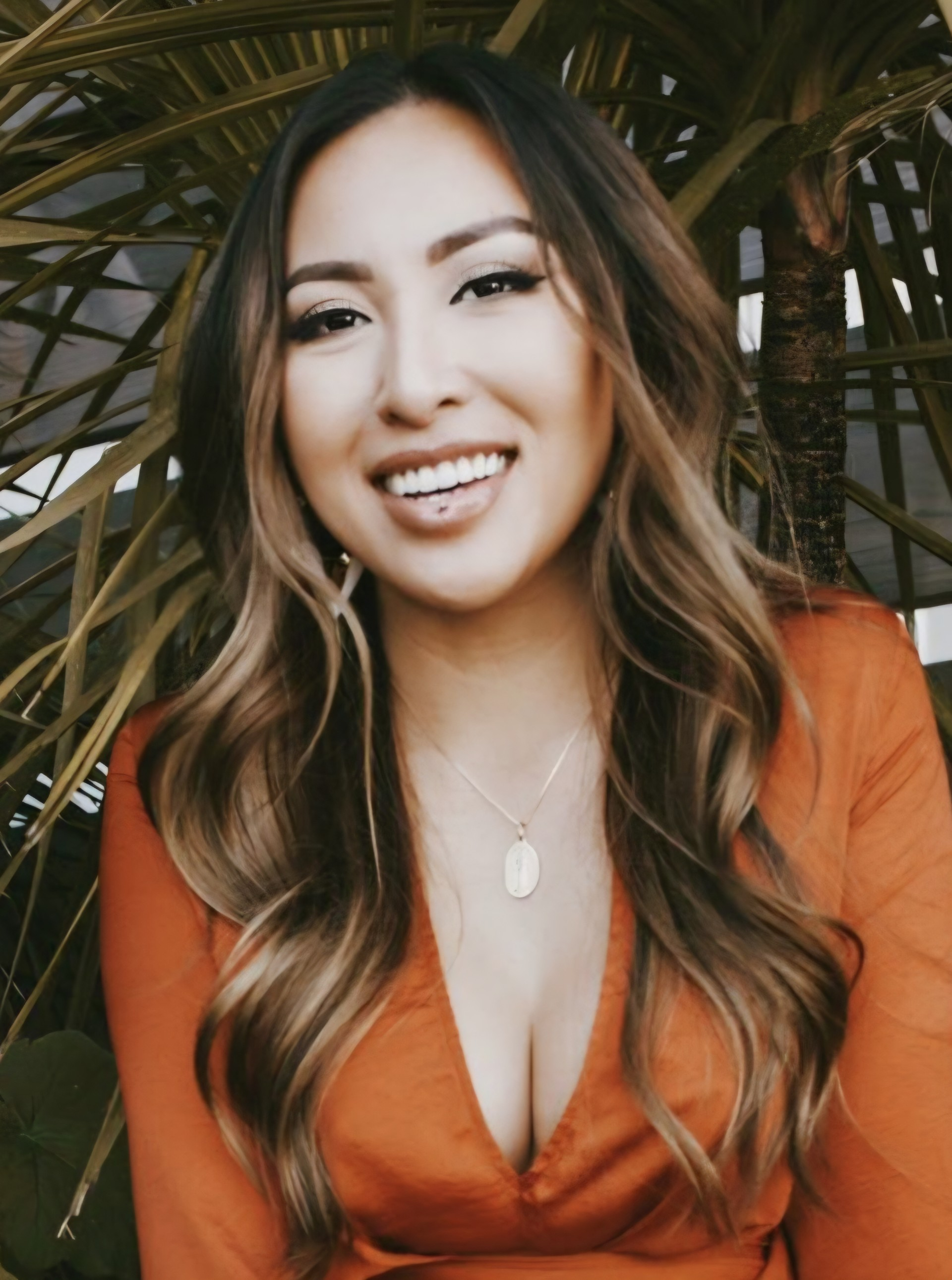 Woman with long, wavy hair, smiling, wearing a black top and necklace, set against a light background.