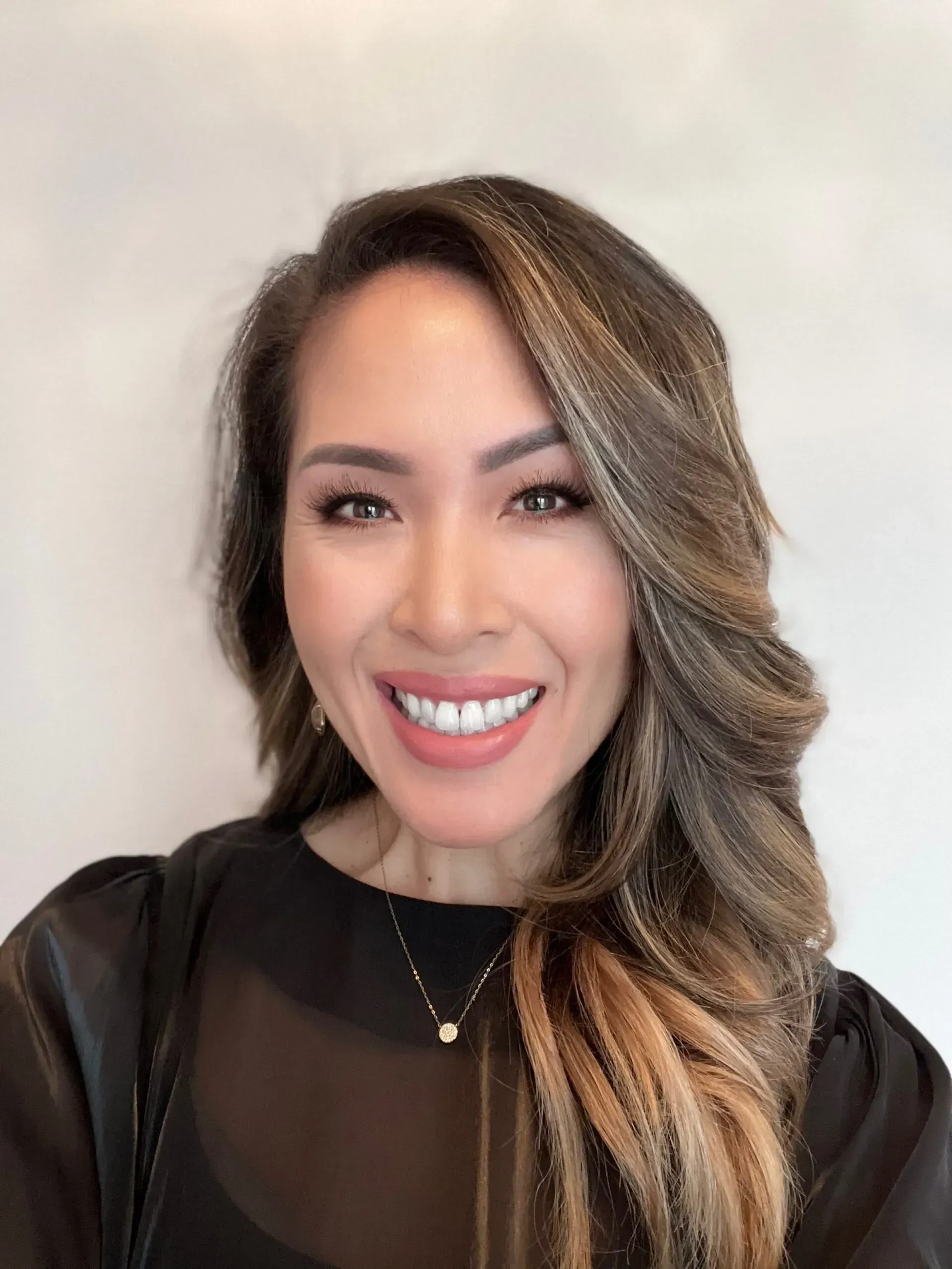 Woman with long, wavy hair, smiling, wearing a black top and necklace, set against a light background.