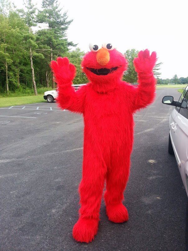 Elmo mascot with arms raised in paved area with trees and vehicles.