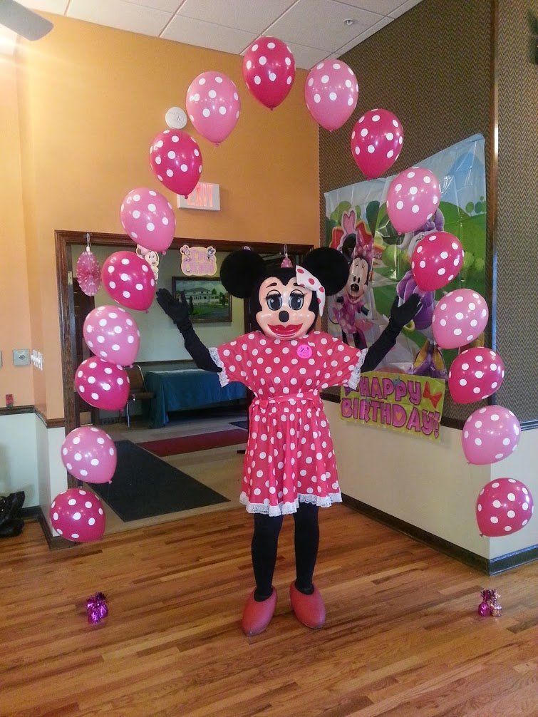 Minnie Mouse costume posing under pink polka dot balloons, arms raised, celebrating a birthday.