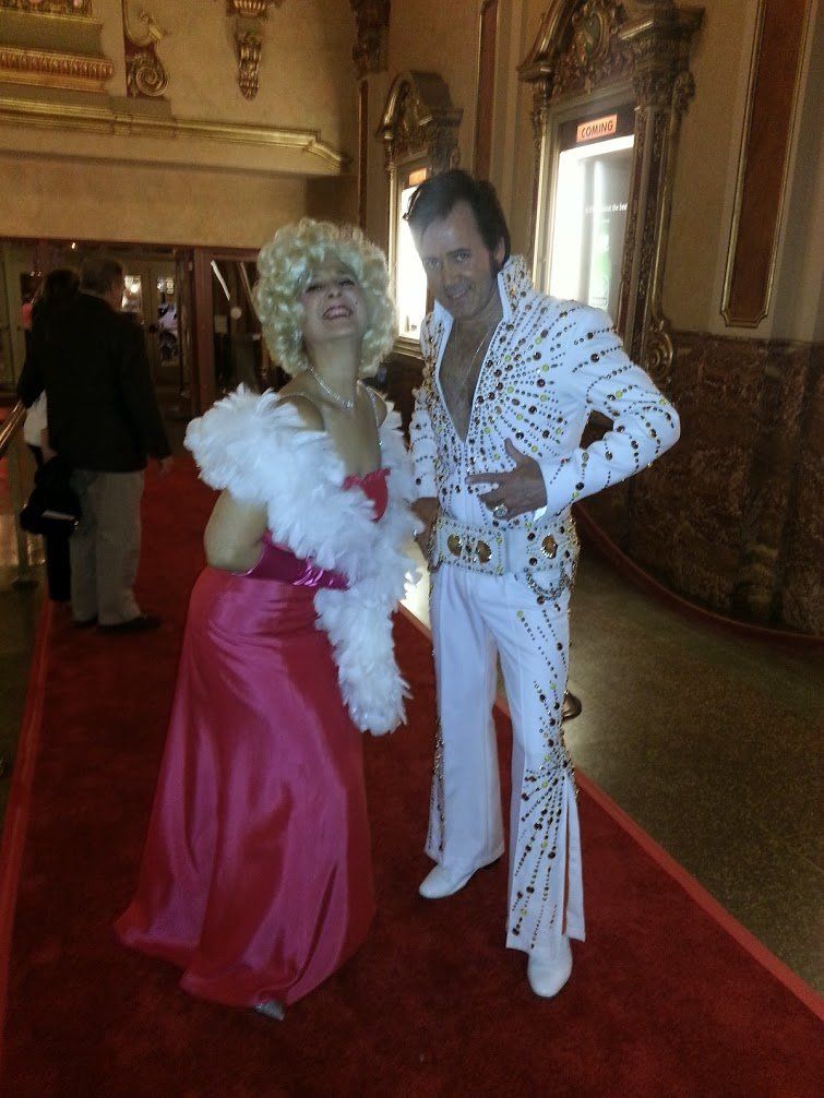 A person dressed as Elvis Presley stands with a woman in a pink gown on a red carpet.