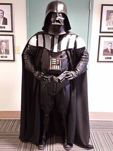 Darth Vader in costume, standing indoors, hands clasped, wearing a helmet and black cape, with portraits on the wall behind.