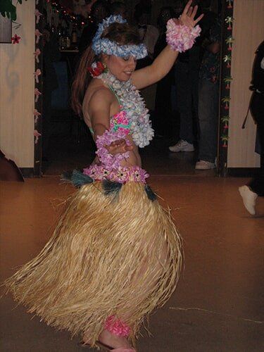 Woman in grass skirt dancing, wearing flower lei, bracelets, and headband. Interior shot.