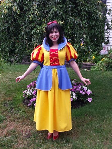 Woman in Snow White costume smiles, arms outstretched, standing in a garden with flowers and greenery.