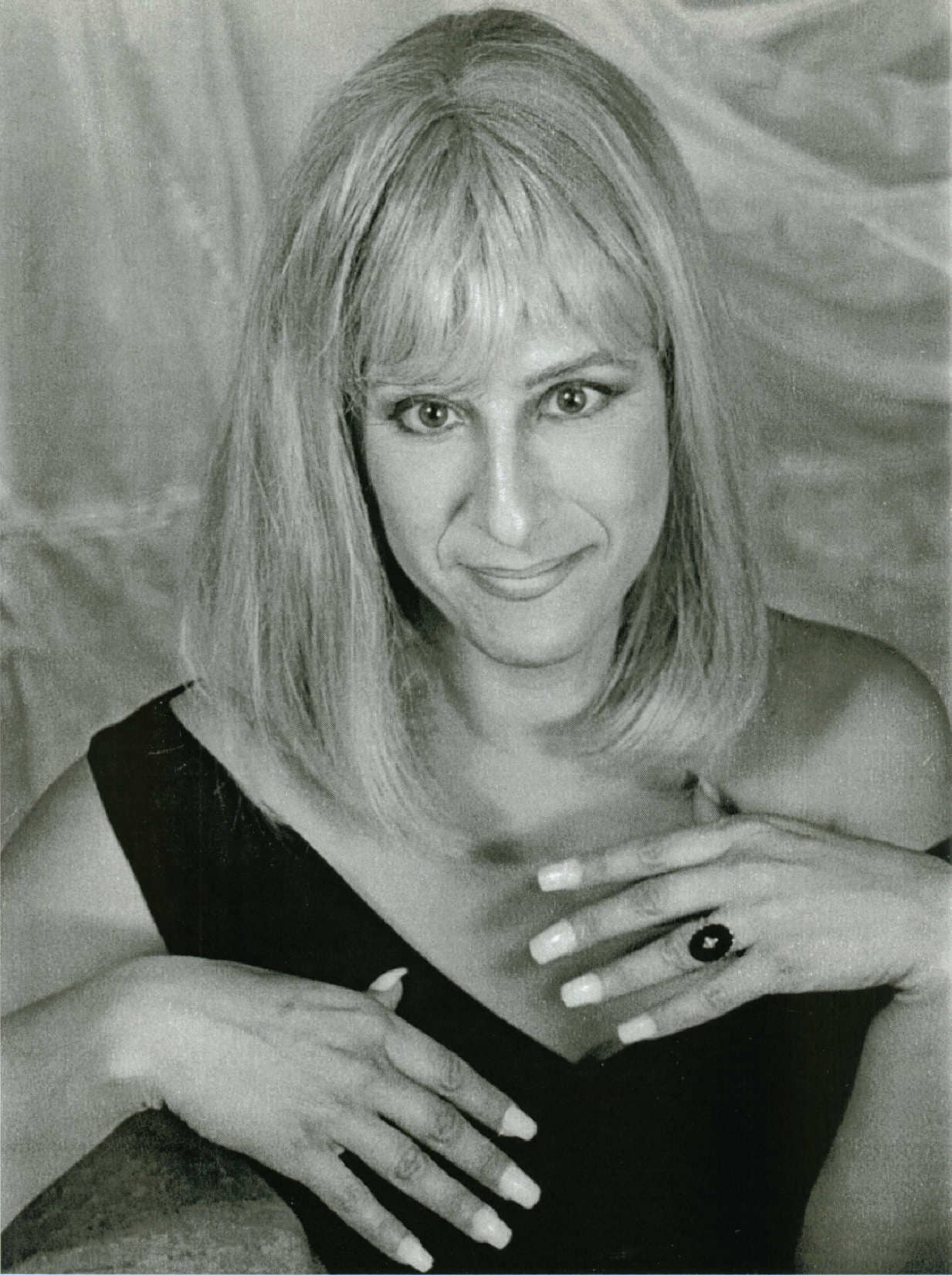 Woman with blonde hair, hands on chest, wearing a black dress and a ring.