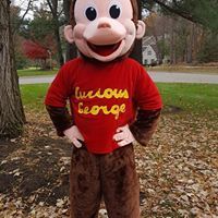 Curious George mascot in red shirt and brown pants standing outdoors in autumn setting.