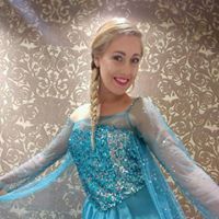Woman in Elsa costume smiles with arms outstretched, against decorative wallpaper.