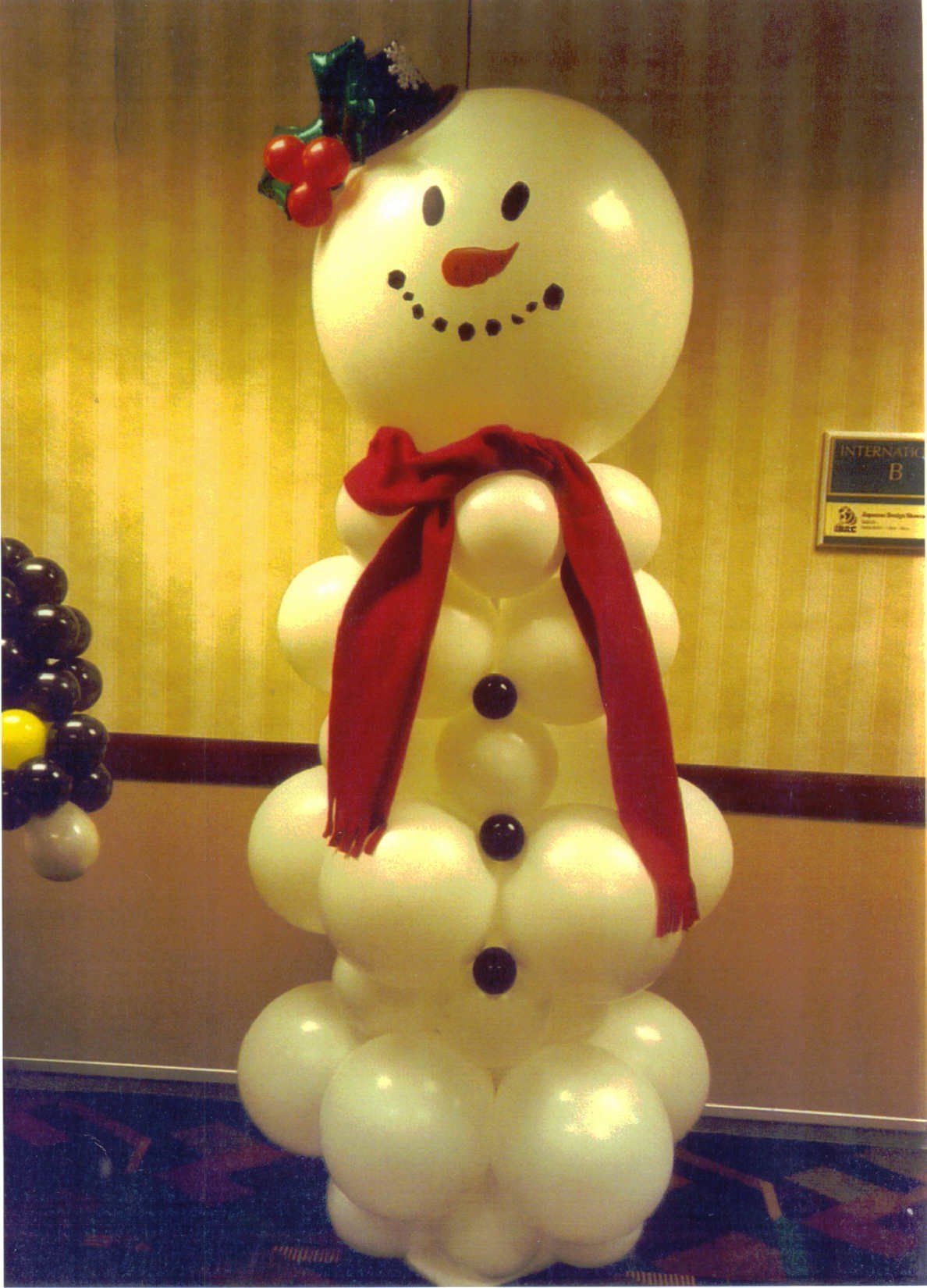 Balloon snowman with red scarf, black coal buttons, and holly on top, set indoors.