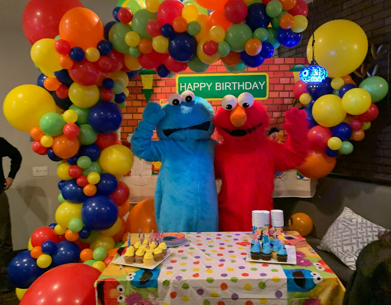Cookie Monster and Elmo stand by a table at a birthday party, with colorful balloons and a 