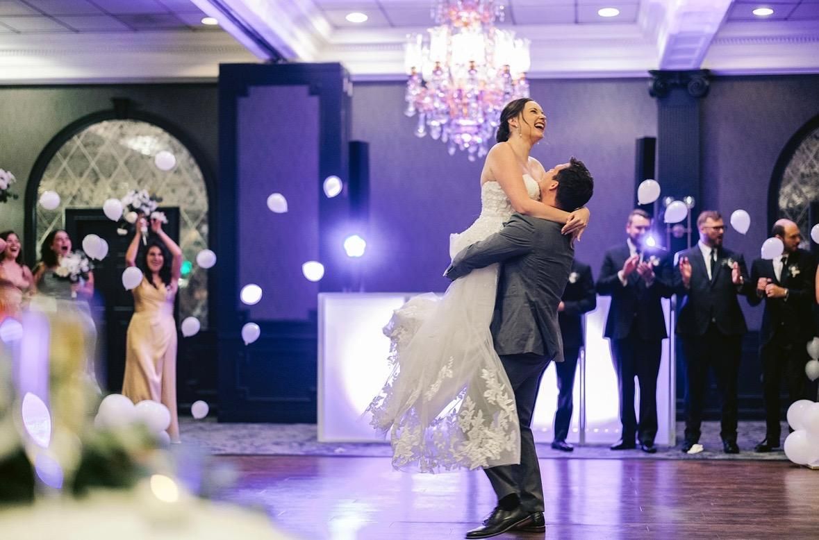 Groom lifting bride in a celebratory pose on a dance floor, balloons in the background.
