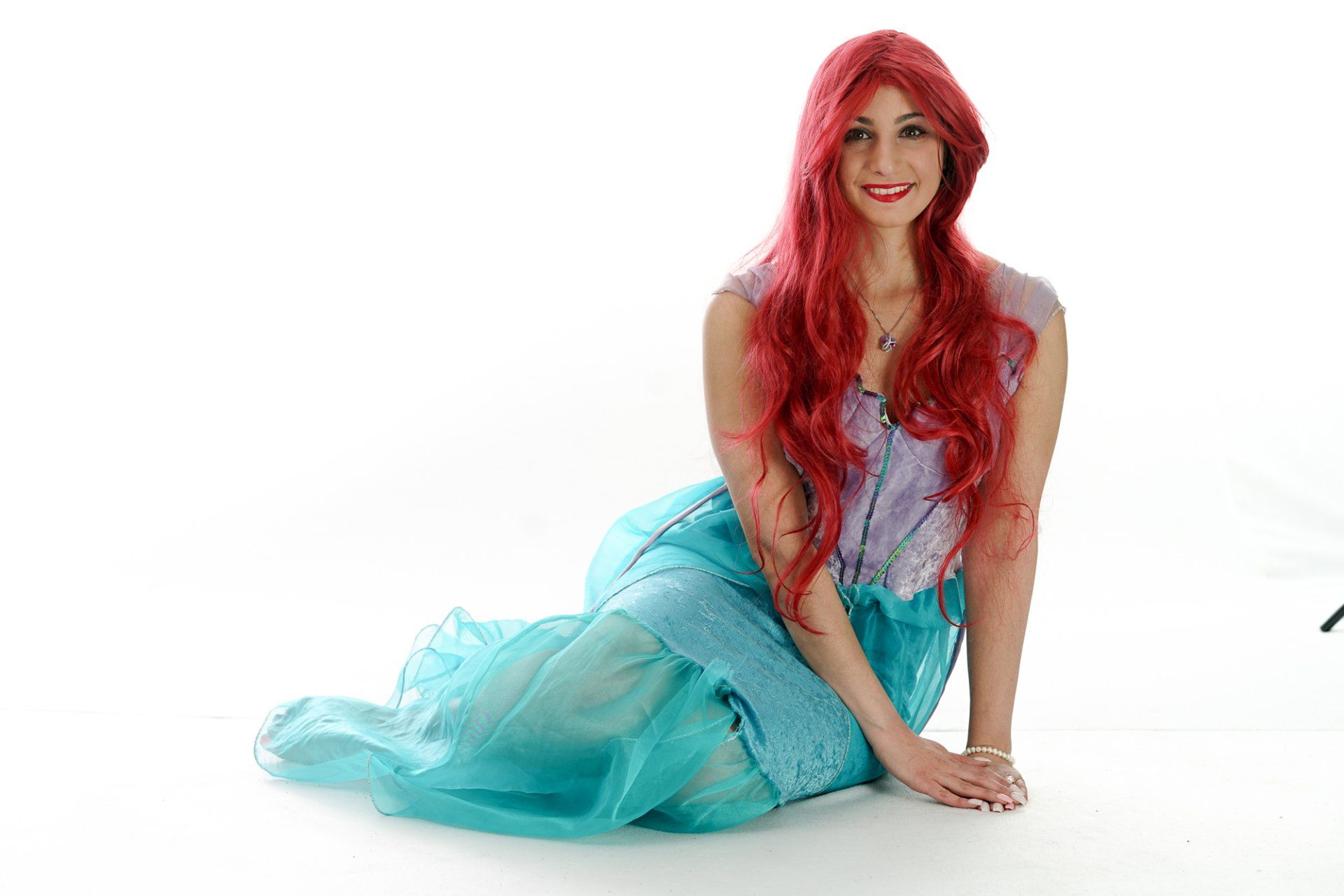 Woman in Ariel costume, red hair, turquoise skirt, sitting on white backdrop, smiling.