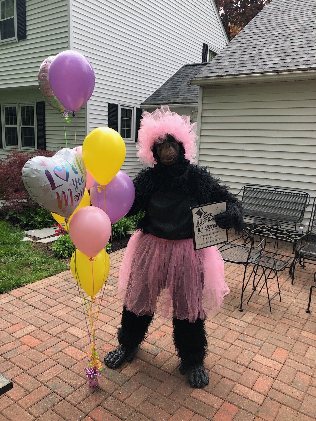 Person in a black gorilla costume with pink tutu holding balloons, standing on a patio.
