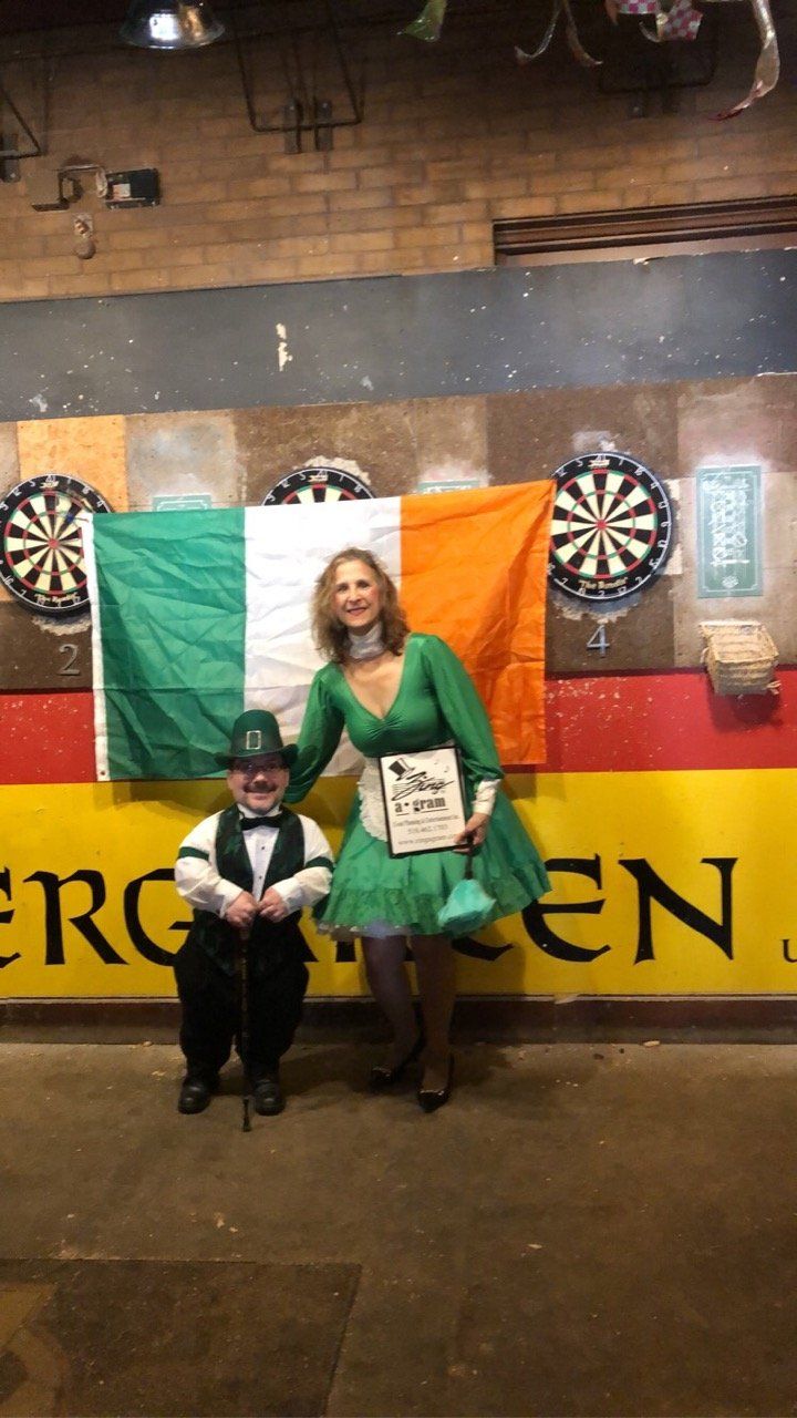 Person in green dress and small person in suit pose near Irish flag with dartboards.