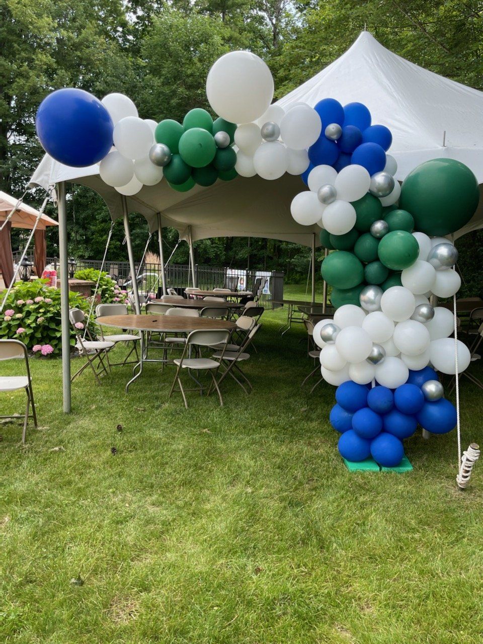 Balloon arch in blue, green, white, and silver, over a tent entrance. Outdoor setting.