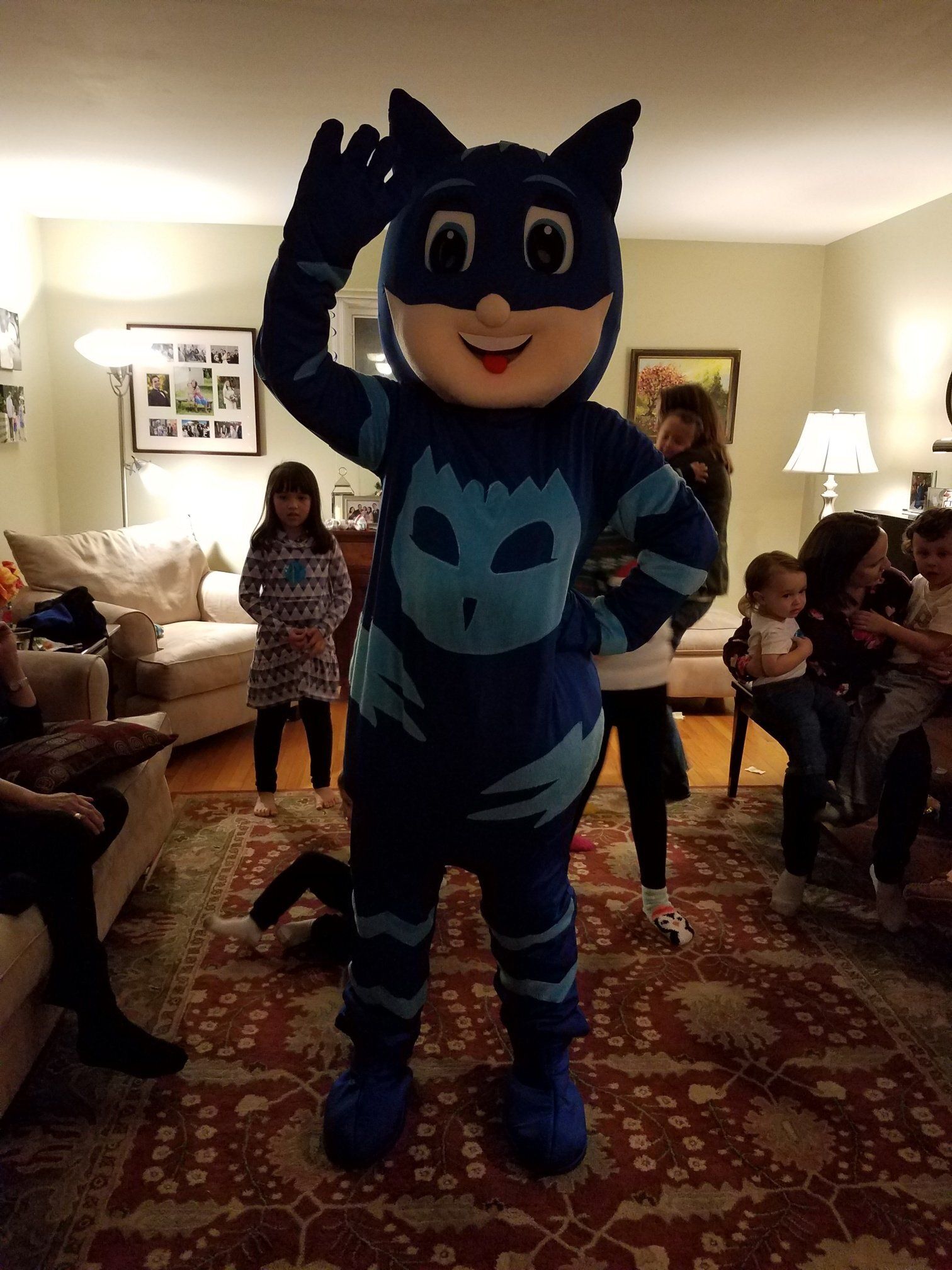 A person in a blue Catboy costume, waving, stands in a living room with children in the background.