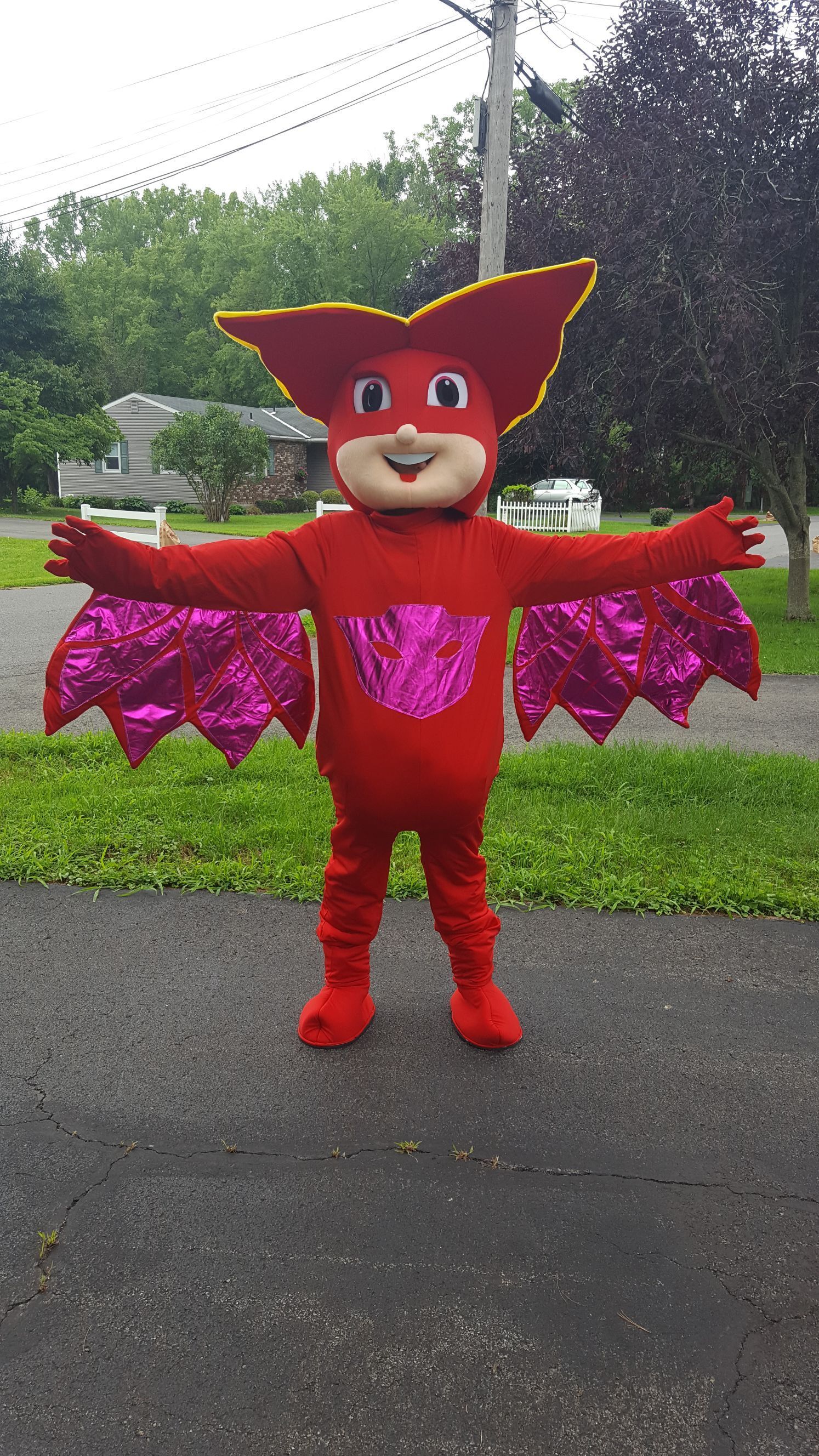 Red bat mascot with pink wings, arms outstretched on pavement in front of green grass and trees.