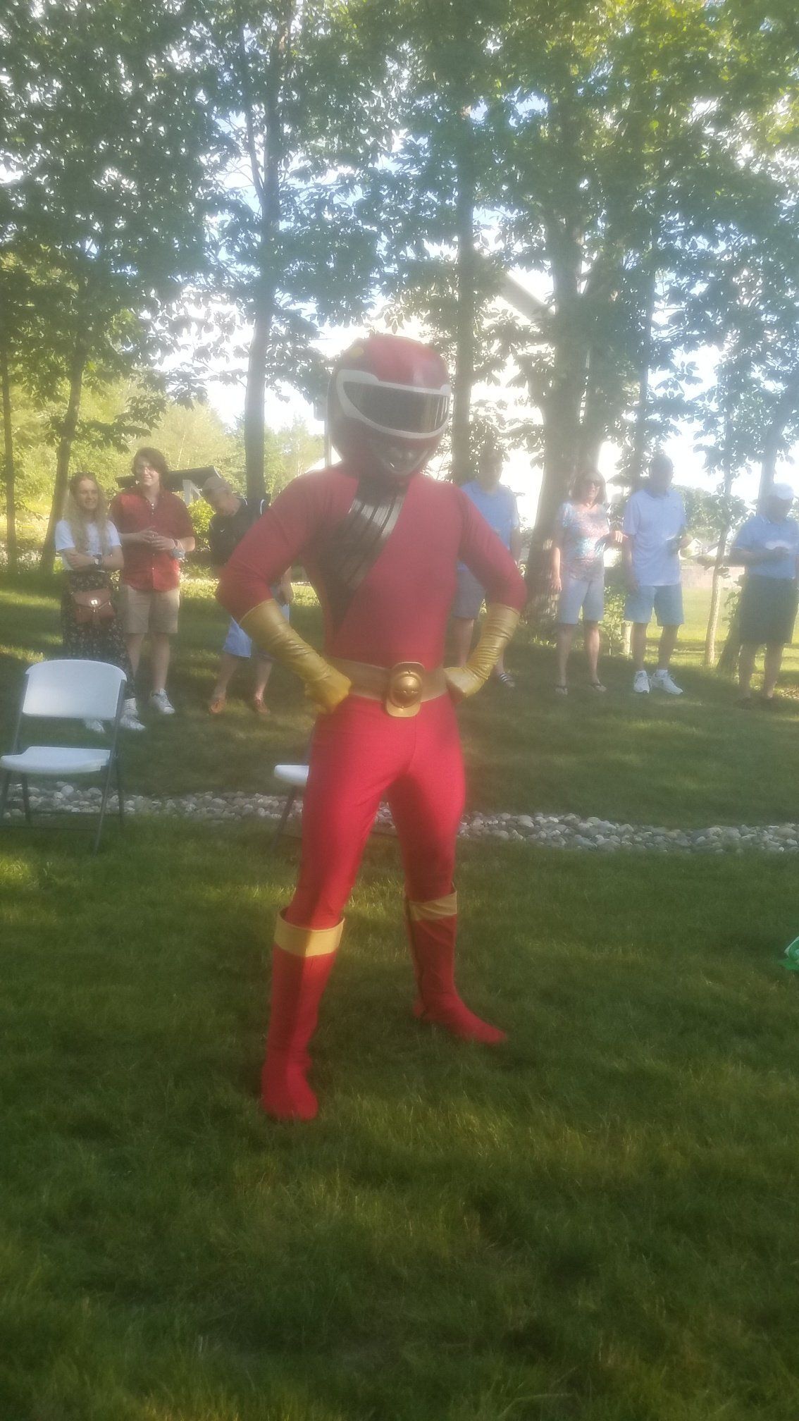 Person in a red Power Ranger costume standing on grass with hands on hips; people in the background.