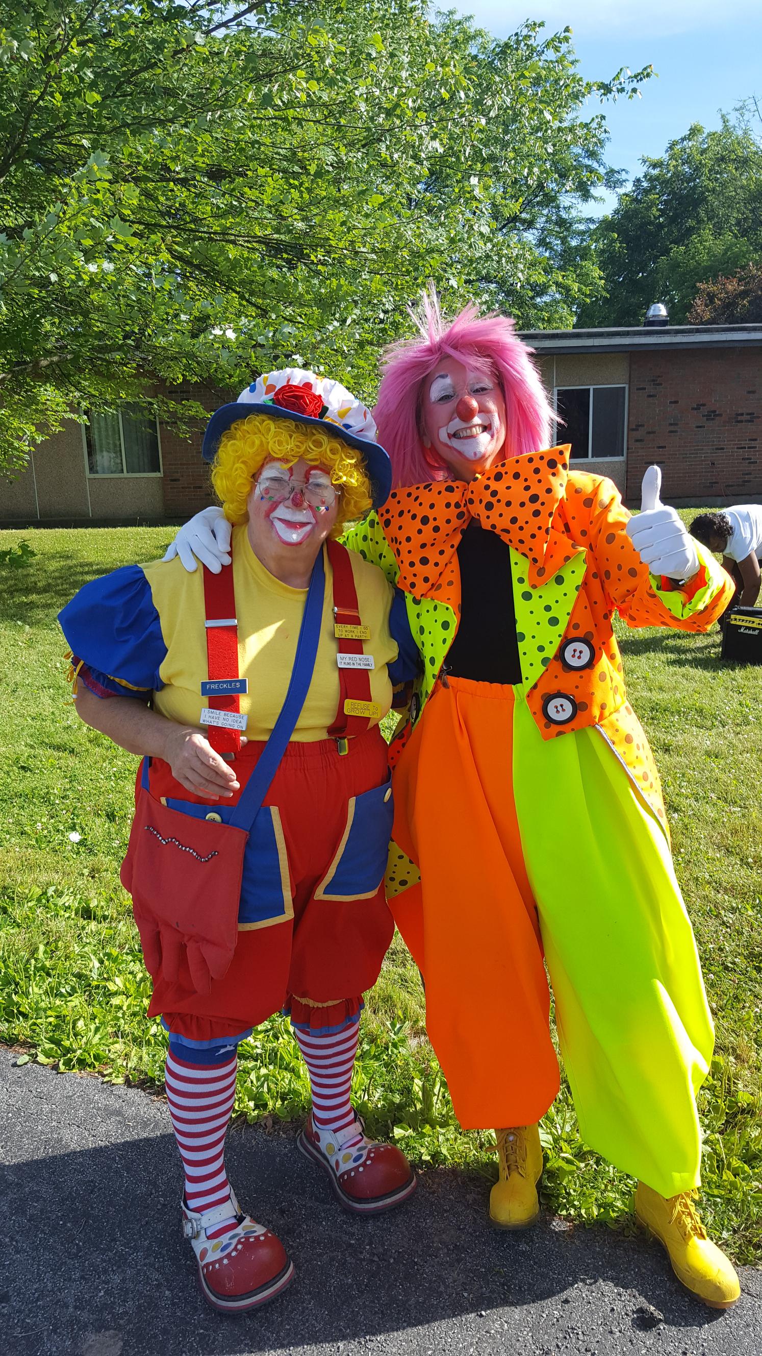 Two clowns in colorful costumes outdoors, smiling. One in yellow, red, blue, the other in orange, pink, and green.