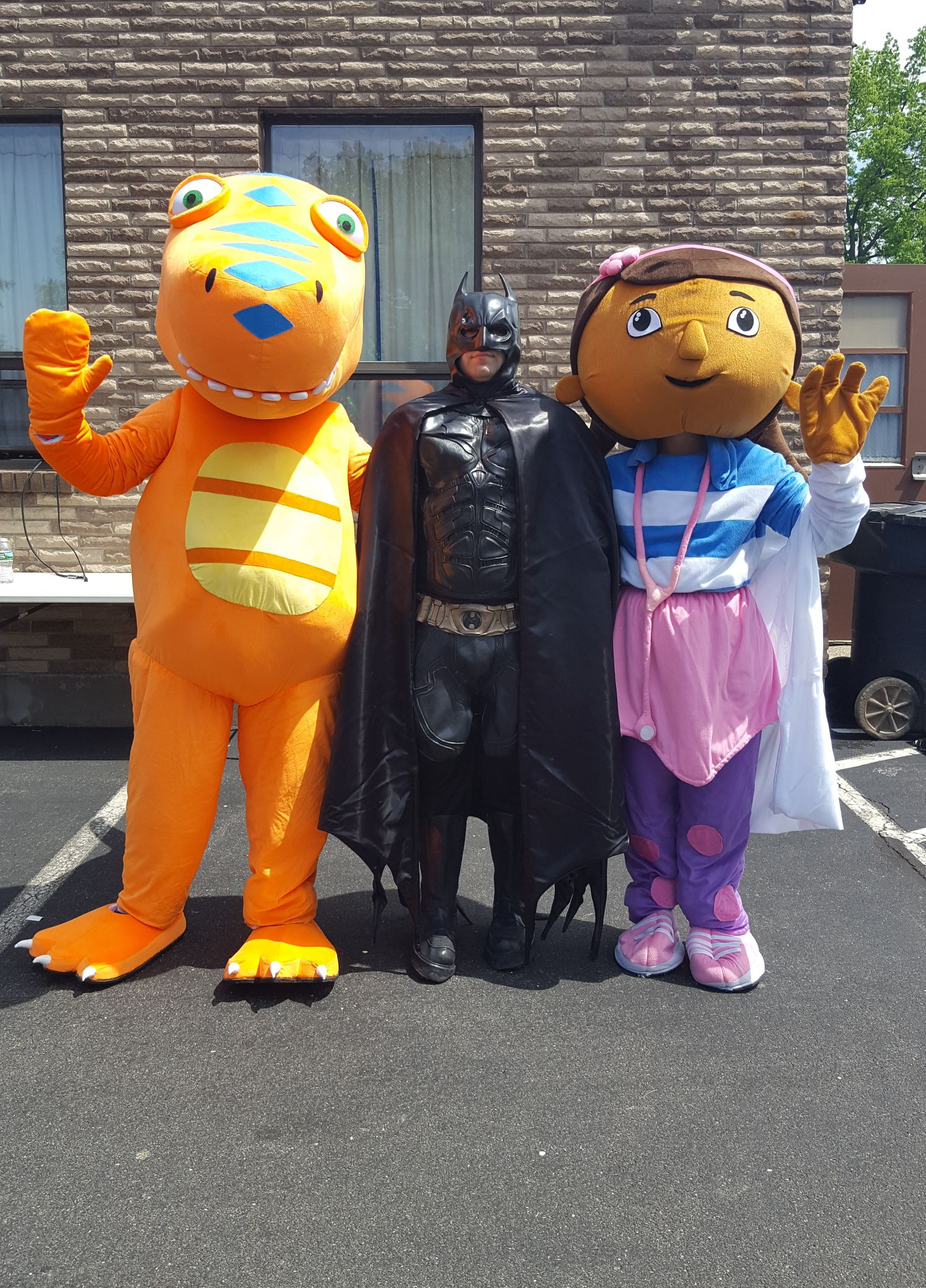 Costumed characters: an orange frog, Batman, and a girl in a purple jumpsuit and pink skirt, standing outdoors.