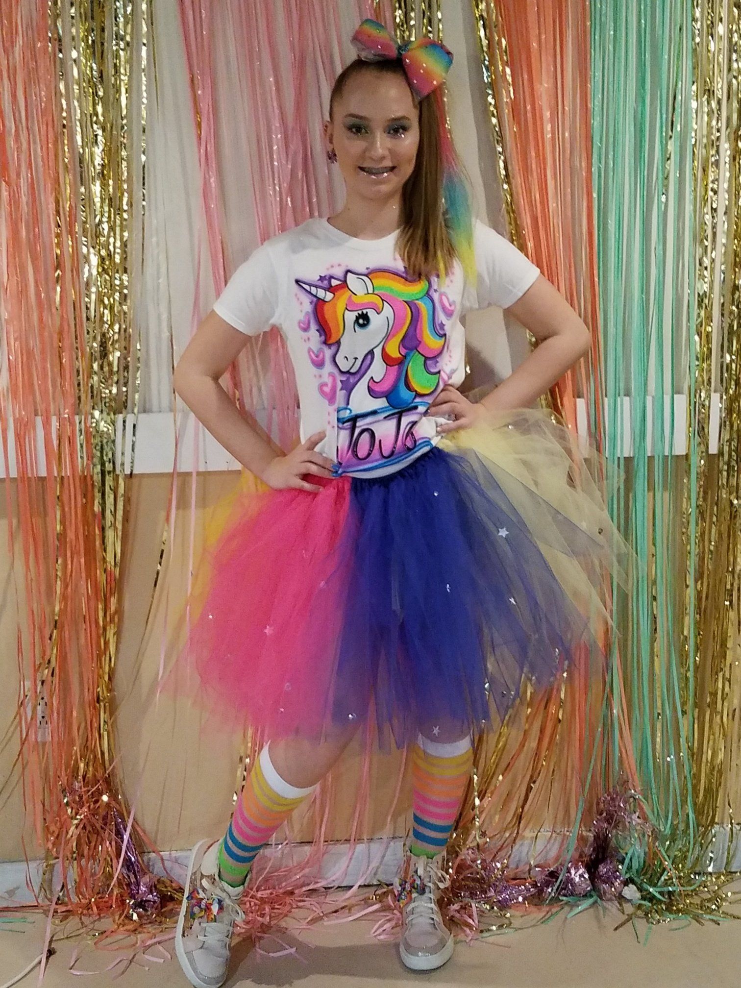 Person in unicorn-themed outfit: white shirt, rainbow tutu, bow, socks, and shoes. Posing in front of colorful streamers.