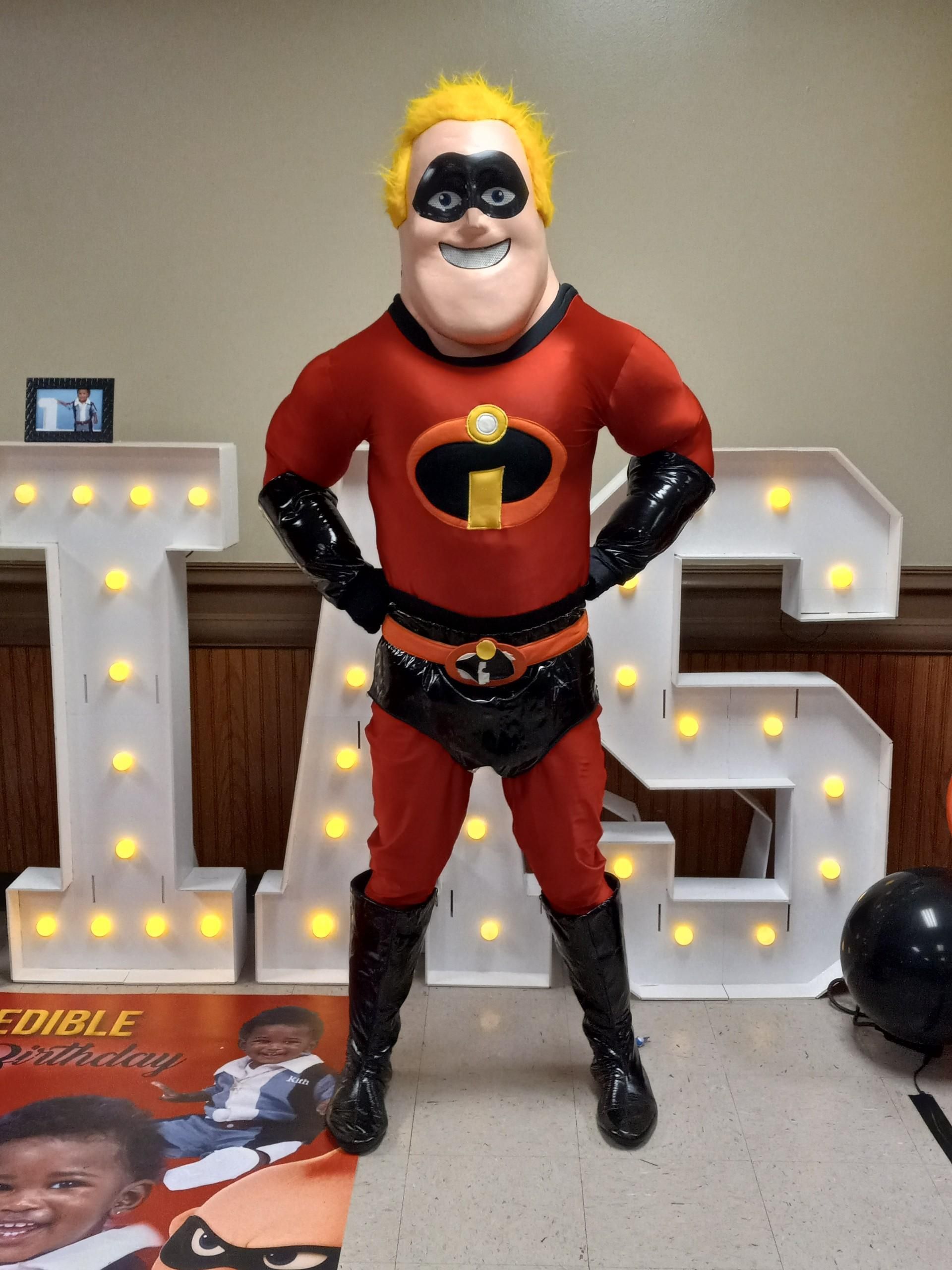 Costumed Mr. Incredible at a party, in a red and black suit with hands on hips, standing in front of light-up letters.